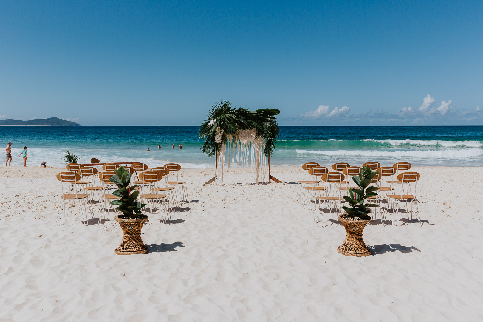 A bright white beach with a deep blue sky/ On the beach is a wedding set up. An arbour, tropical florals and wooden chairs