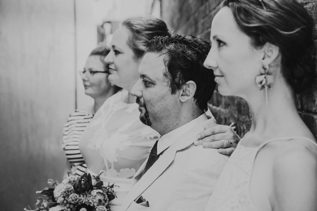 Four people stand in a line against a wall. The image is shot from the side, viewing them all in profile. It looks like a band's album cover but it is a wedding photograph