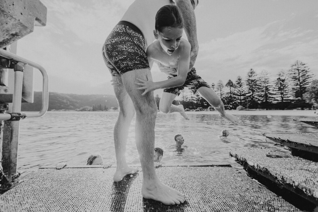 Black and white image of a young boy being pulled from the ocean by his father on patonga Wharf during a real-life family photography session