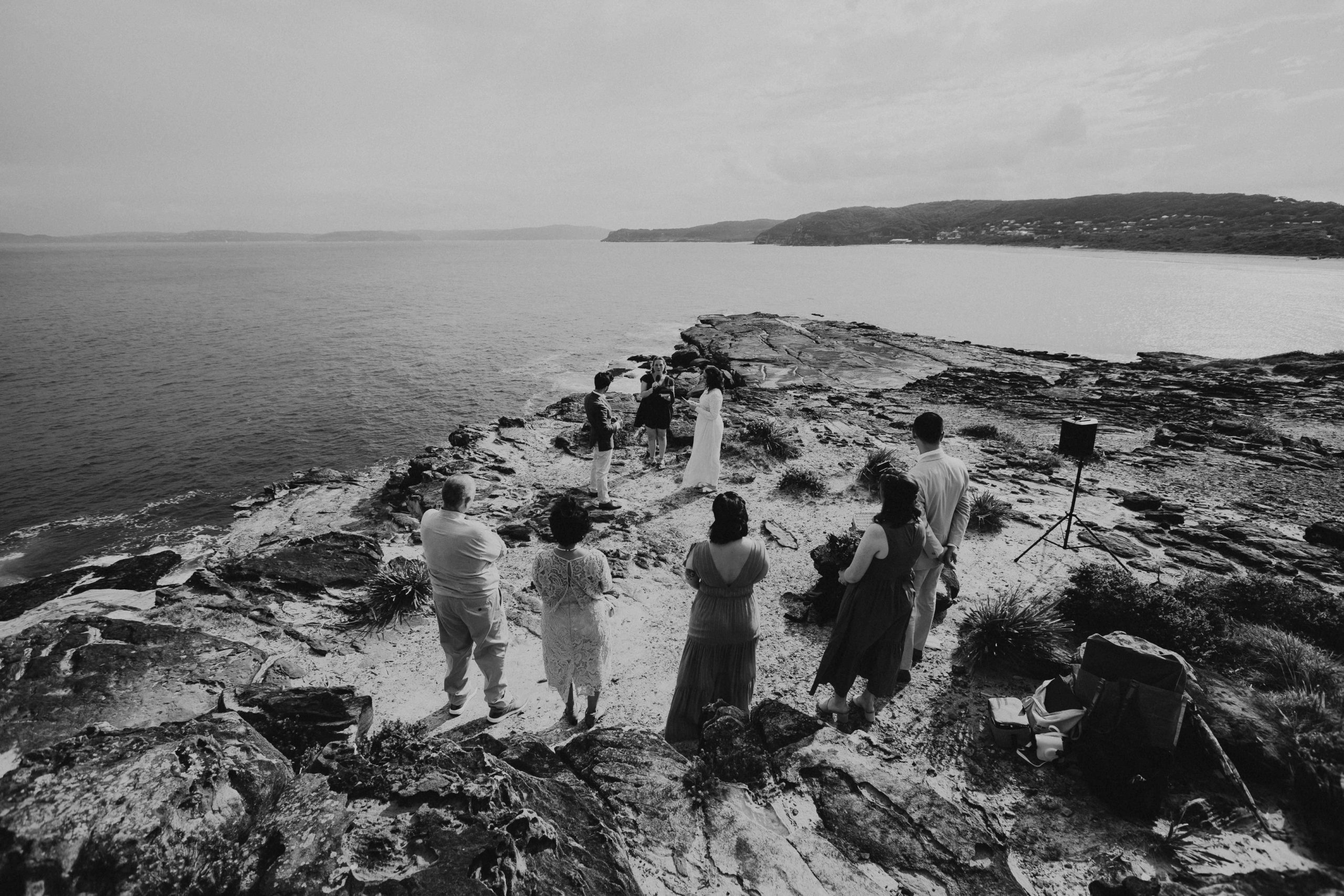 A small group of people watch on an Elopement takes place on a clifftop in Bouddi NP on the Central Coast. Black and white image