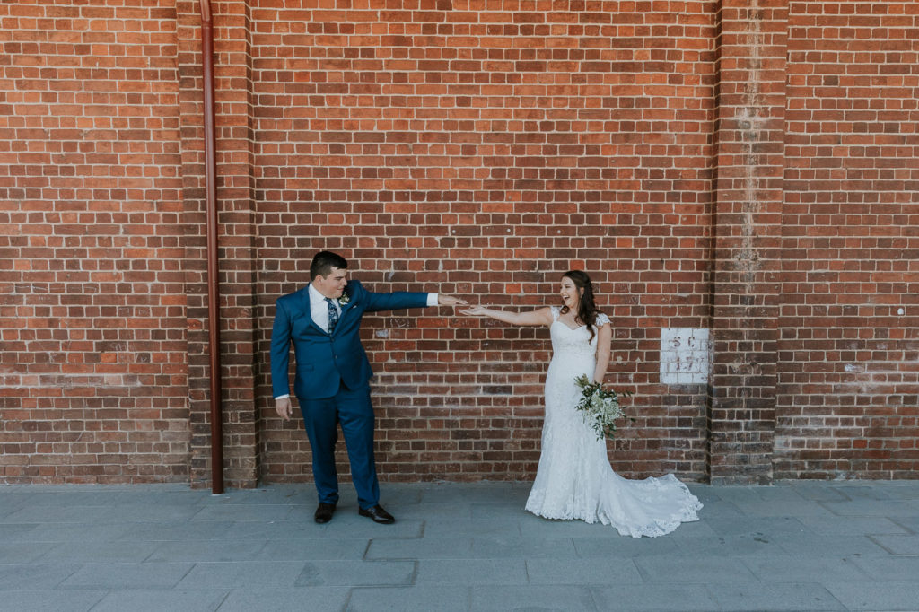 Image of a young married couple standing in front of a brick wall. They stand apart, jokingly observing the new Covid restrictions of social distancing but holding hand from afar. It is fun. Wedding shot at Wodonga Junction Square.