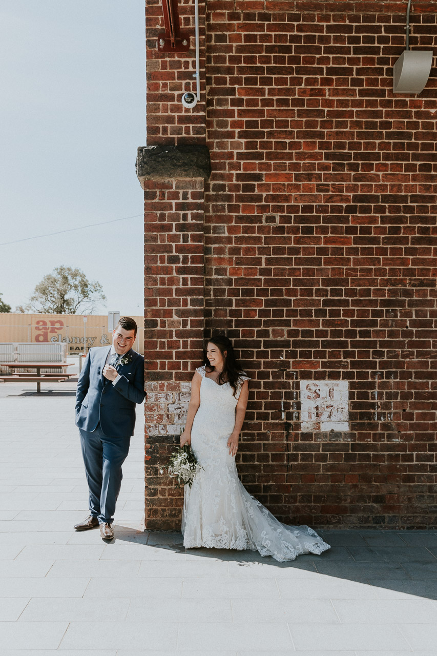 Bride and Groom casually lean against old brick wall, laughing towards each other
