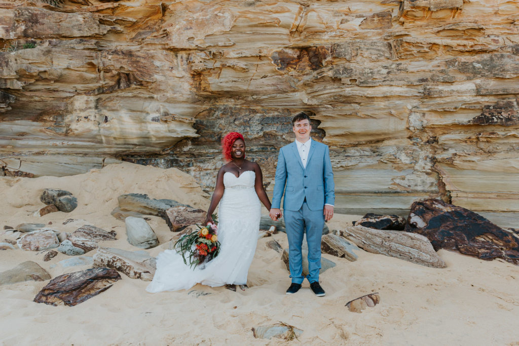 A black african lady wears a bright white wedding dress and bright red hair. She is holding hands with her white husband, who wears a light blue suit. They stand in front of a sandstone cliff on MacMasters Beach, Central Coast NSW