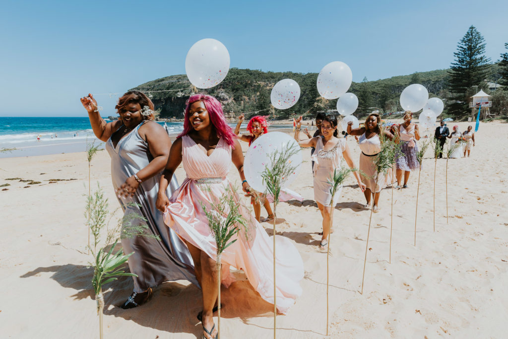 A procession of African ladies, walk down the beach singing and chanting, and carrying white balloons. It is a Central Coast Wedding on MacMasters Beach. Very fun image.