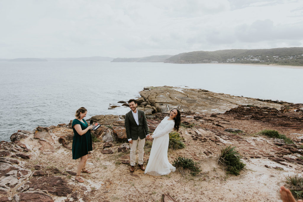 A couple get married in their Elopement with a Celebrant standing close by. They are standing on rock with an amazing vista behind them on the NSW Central Coast. They are laughing and having fun