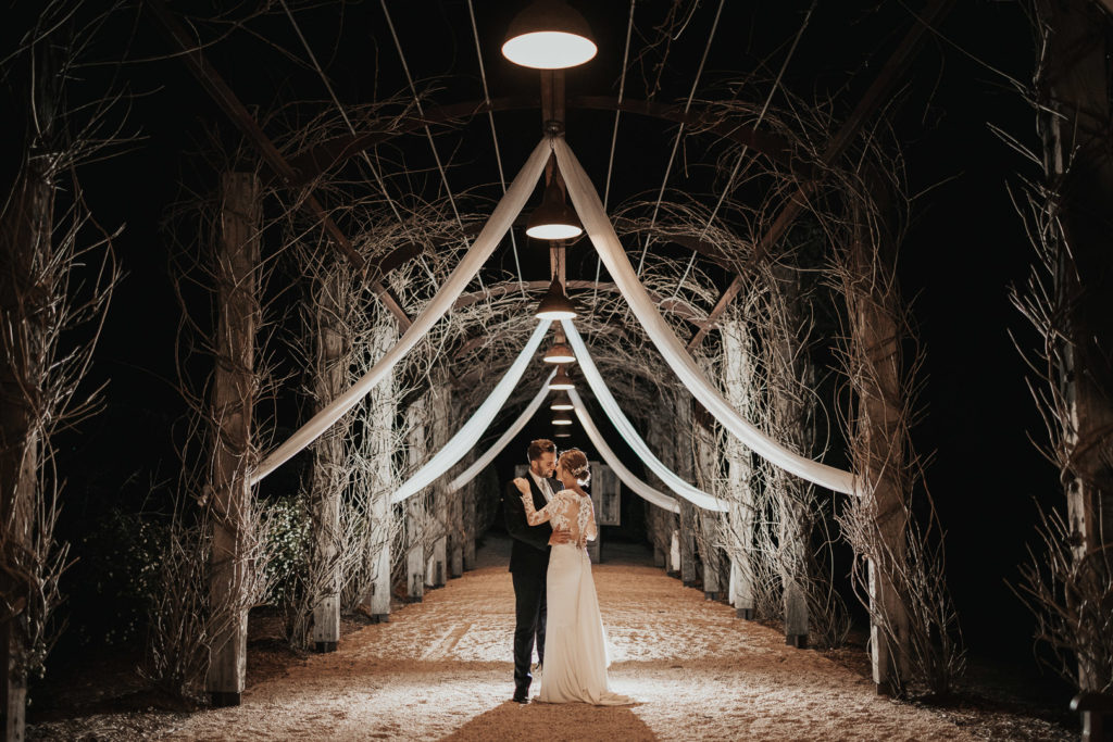 Night photograph of a couple standing under a huge pergola. They are backlit. The image is romantic and dramatic