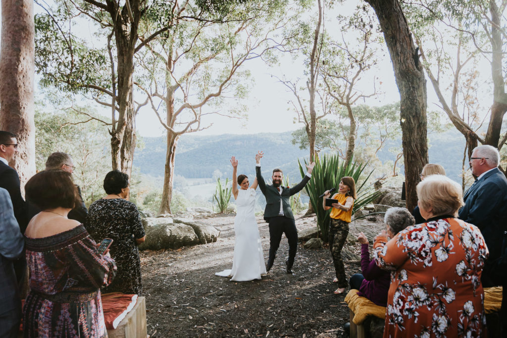 In a wedding at Glenworth Valley, the bride and groom celebrate with arms in the air. They are surrounded by trees and woodland with a lookout behind them.