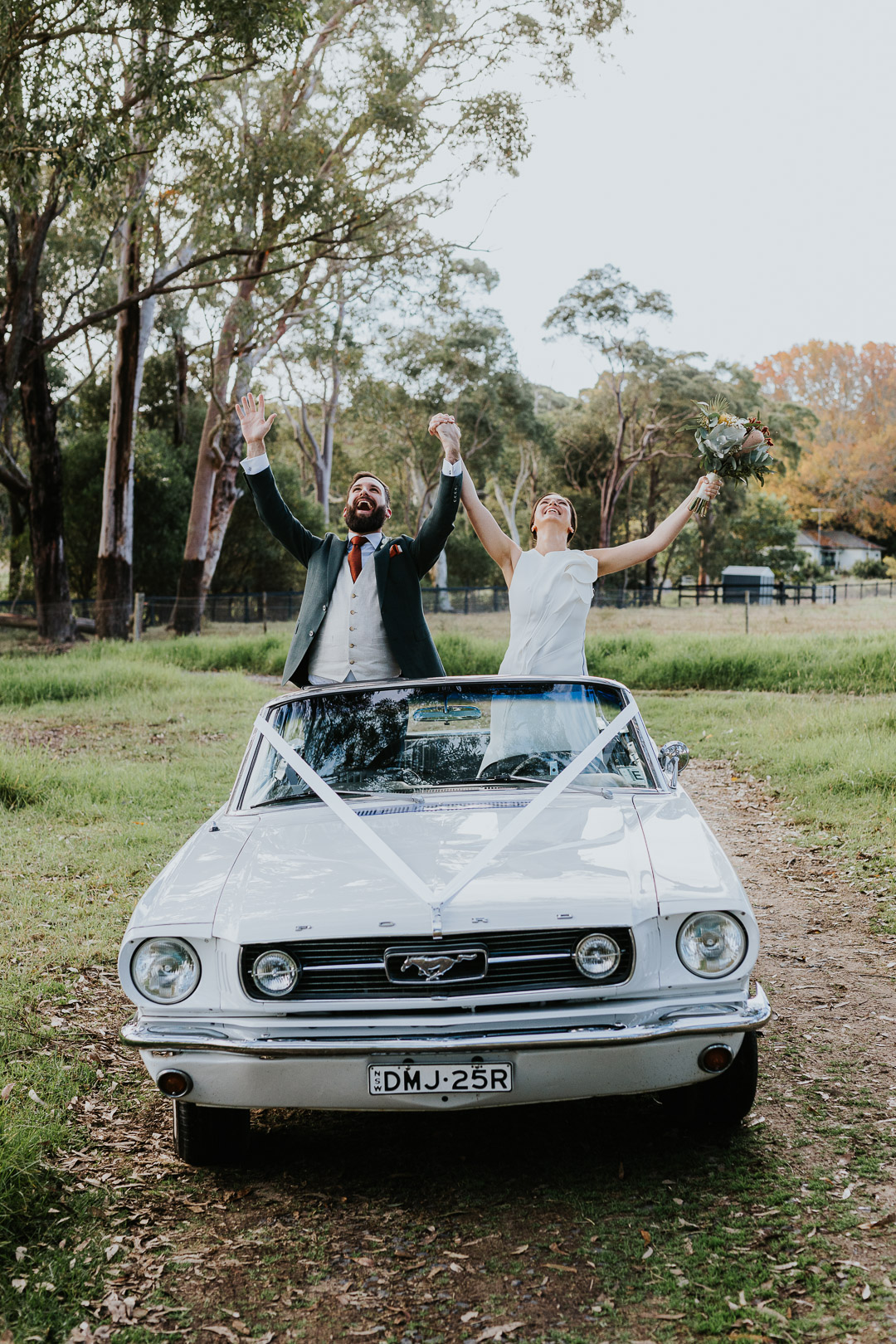 A couple stand up in an open-top Convertible Mustang car. They are in the countryside. They throw their arms up in celebration. They just got married. Glenworth Valley, NSW Wedding Photographer.