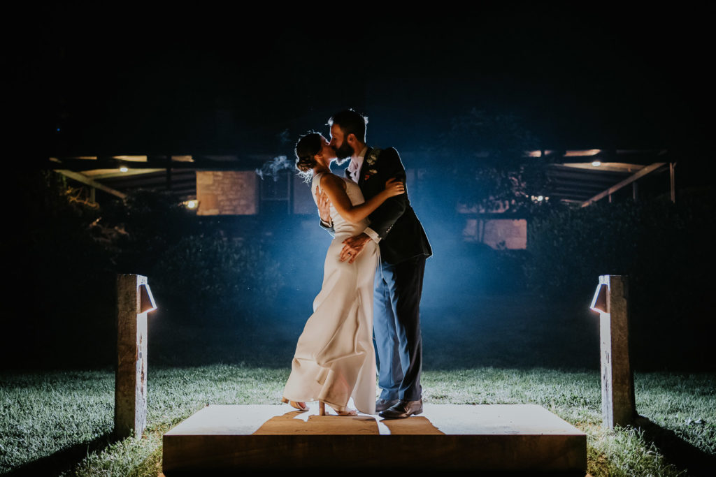 A married man and woman kiss in a dramatic night photo. There is blue light emanating from behind them. They are at an outdoor wedding venue.