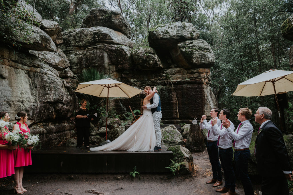 A couple stand in the middle of the picture. They have just got married. They kiss in front of their friends and family. They are in a rock valley at Kangaroo Valley Bush Retreat. It is damp and wet.