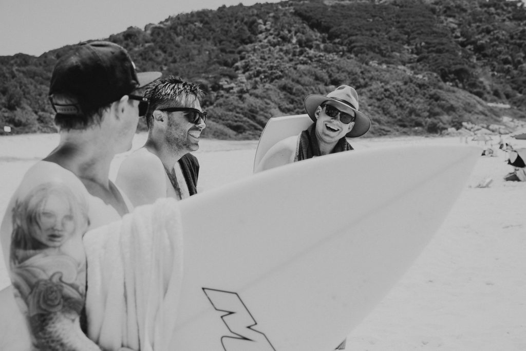 A group of friends carrying surfboards talk and laugh before their wedding at Forster, NSW. Black and white image.