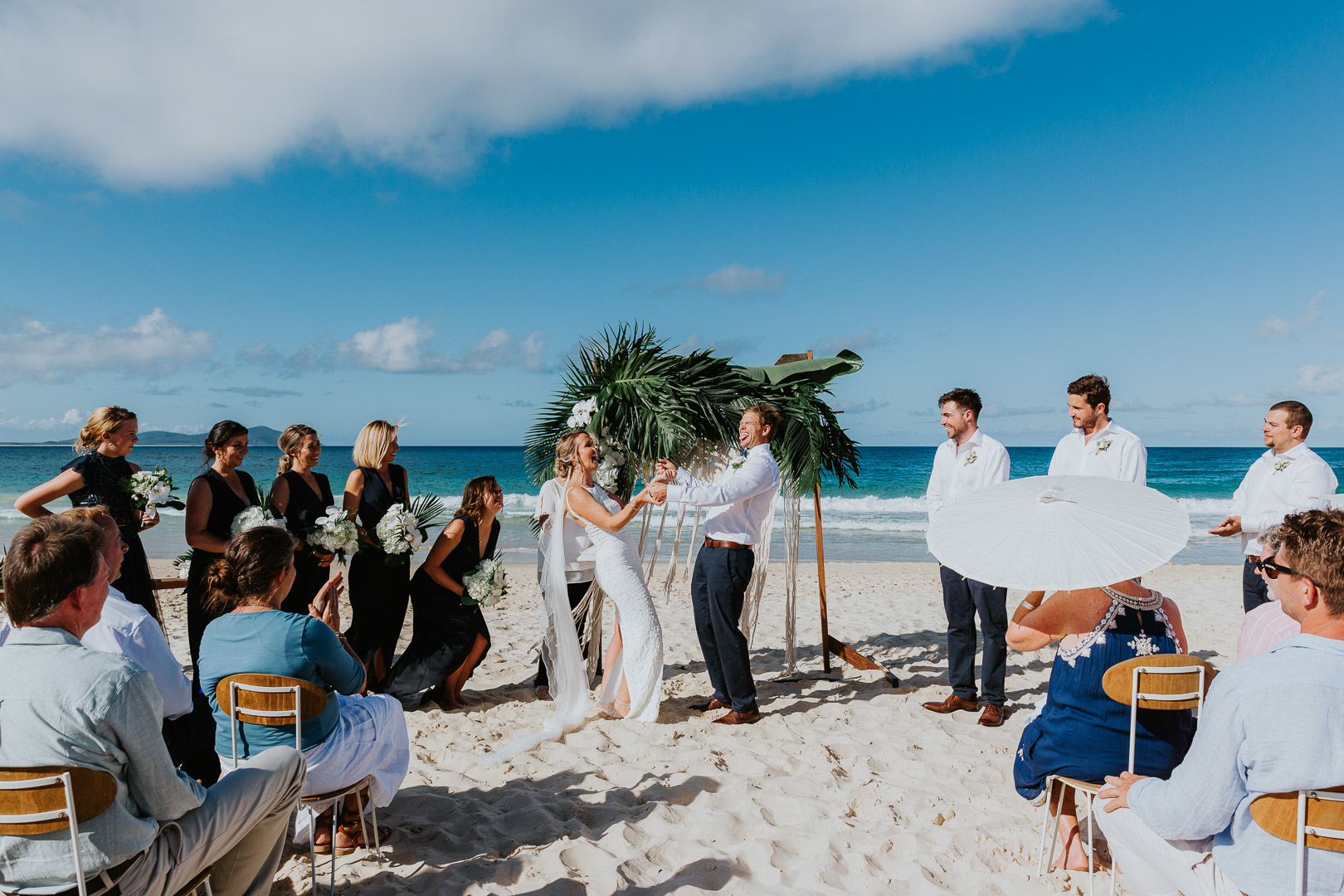 A couple who have just got maried celebrate in front of their Tropical styled arbour on a pristine white beach, with a bright blue sky above. Their guests cheer around them