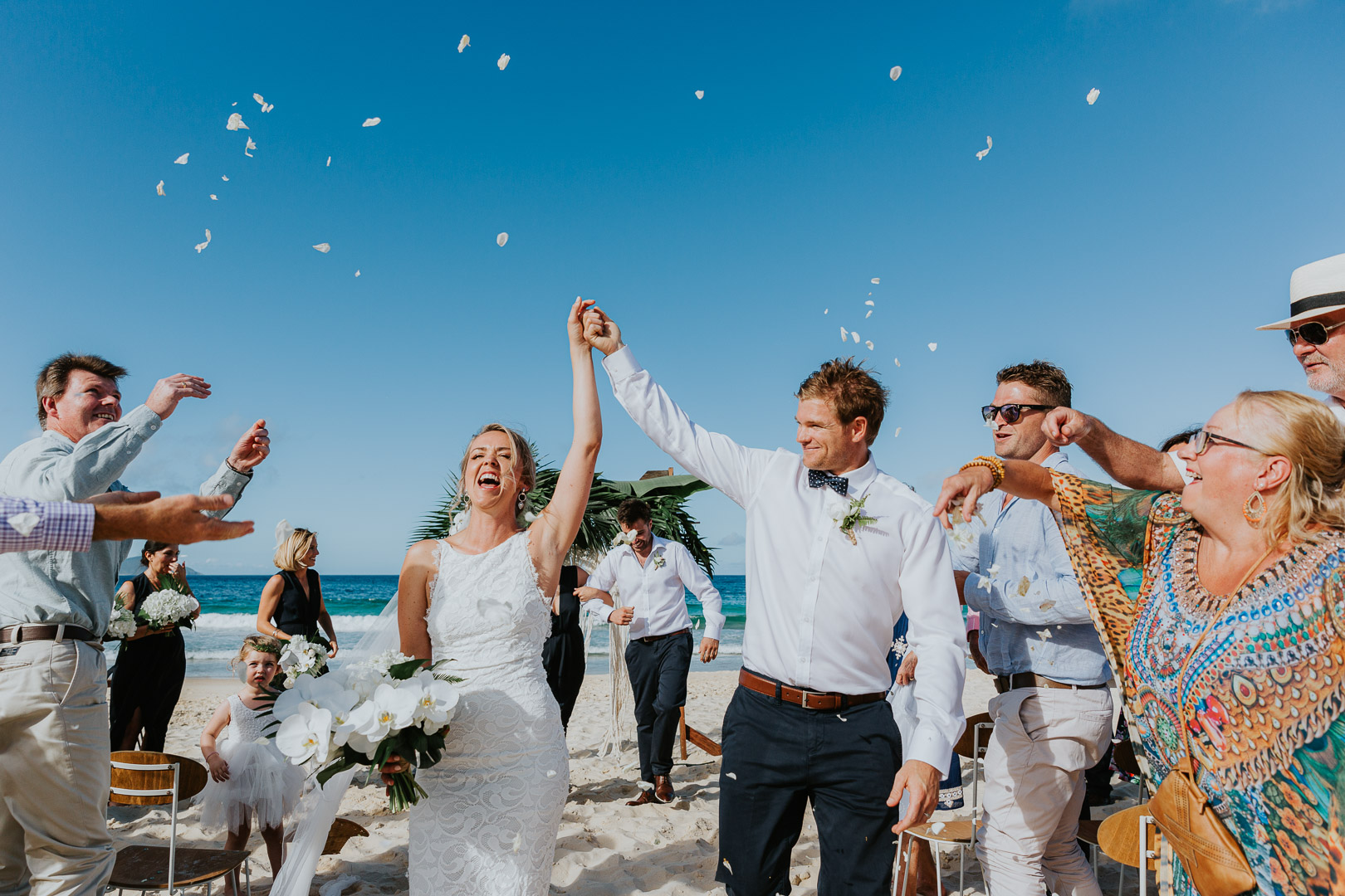 A married couple walk down the aisle on the beach with an enormous bright blue sky behind them whilst their guests throw petal confetti over them. The yell with joy. It a very happy and bold photo.