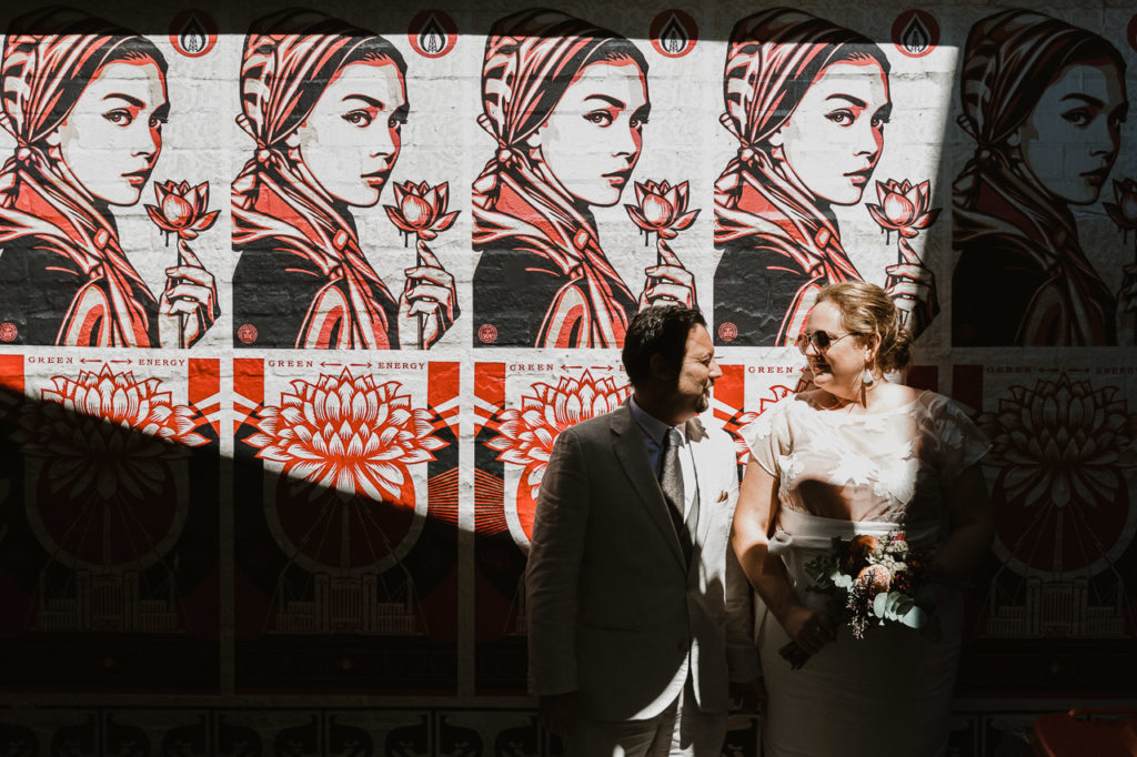 A wall covered in a repeating poster of a woman which is dominated by red. The sun falls dramatically across the wall creating hard shadows. A man and woman look at each other, half in and half out of the shadows.