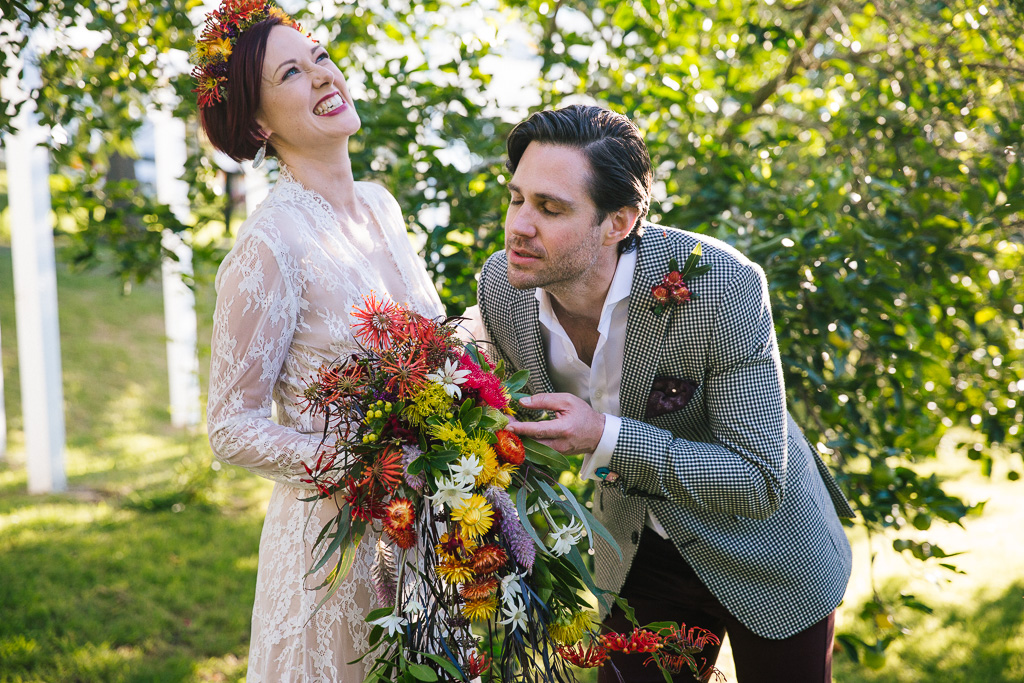 Fun and colourful photo of a groom smelling the flowers in his wife's bouquet. She is laughing. Lots of Australian natives and colour