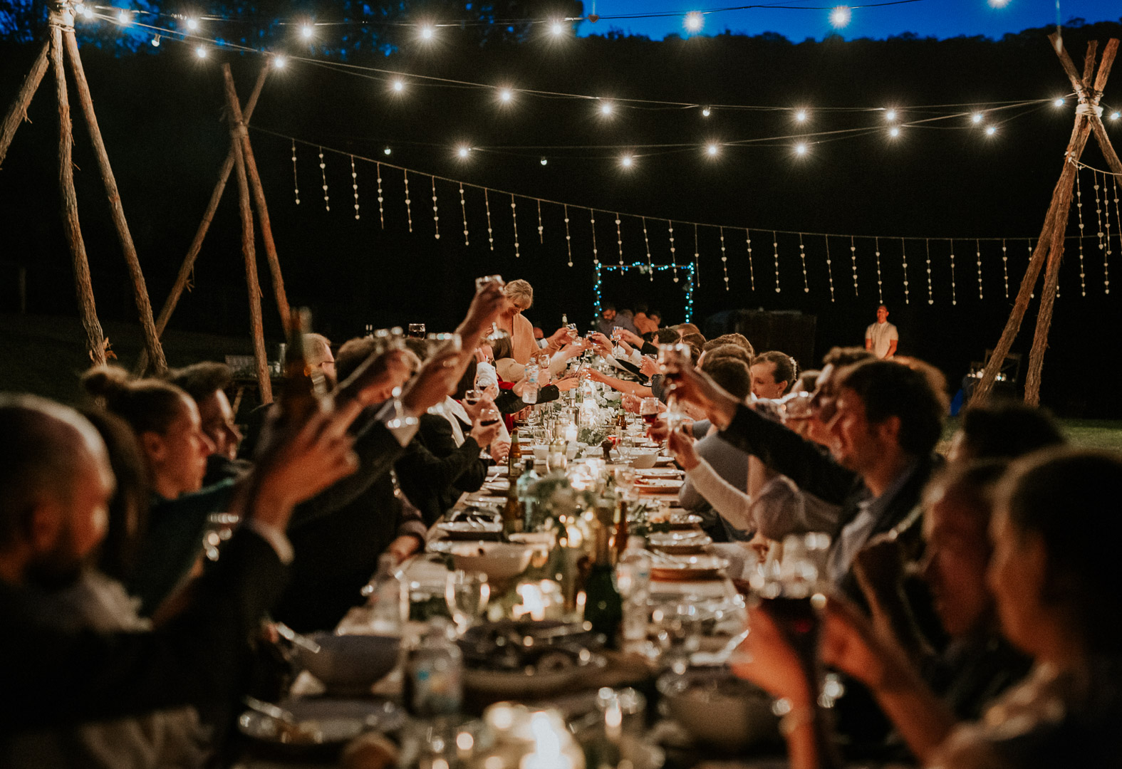 A photograph taken down a long table with guests either side all raising their glasses to cheers. Outdoors under fairy lights
