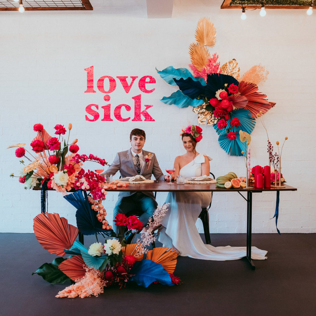 A young couple sit behind a bridal table in a fashion-like image. There is lots of colourful floral and styling and behind them are the words Love Sick