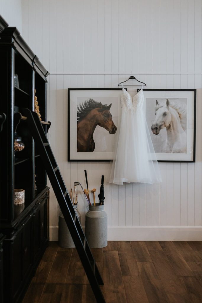 A wedding dress hanging in front of two framed images of horses. The colours of the room are black and white. Dark Horse Vineyard, Hunter Valley.
