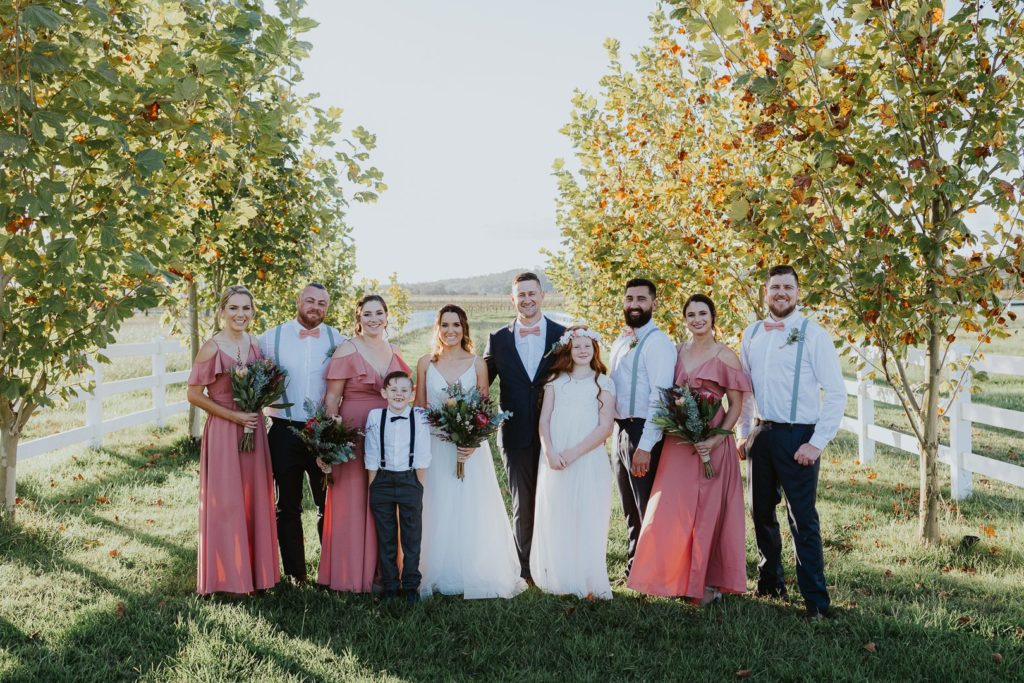 Wedding Party standing in the pear trees at Dark Horse Vineyard in the Hunter Valley. The girls wear a blush pink dress and the boys are in white shirts with light blue braces and pink bow ties. They all look happy and there is beautiful sunlight.