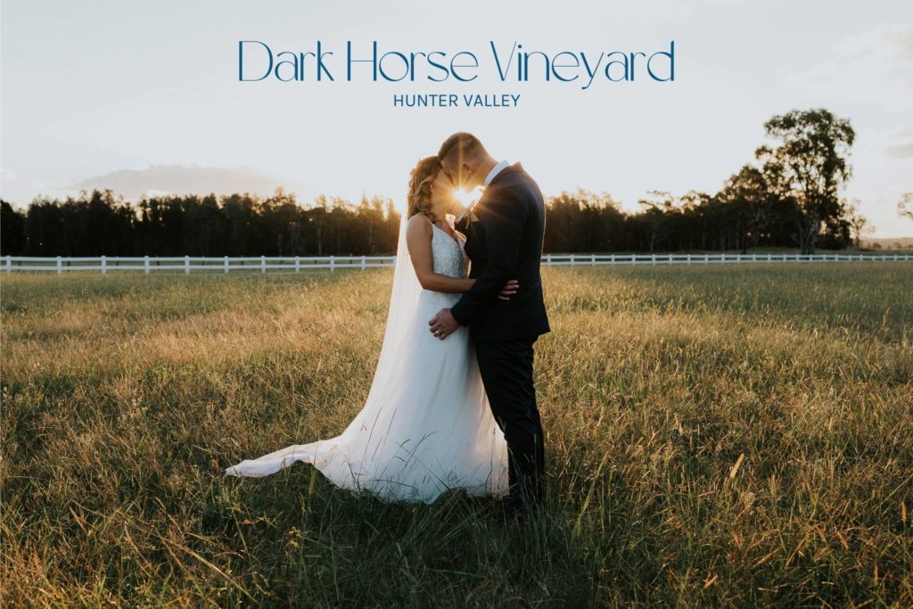 A beautiful photograph of a couple kissing on their wedding day. They stand in a field of long grass with the sun behind them. The gap in between their heads has created a love heart with the sun burst. Dark Horse Vineyard in the Hunter Valley, NSW, Australia