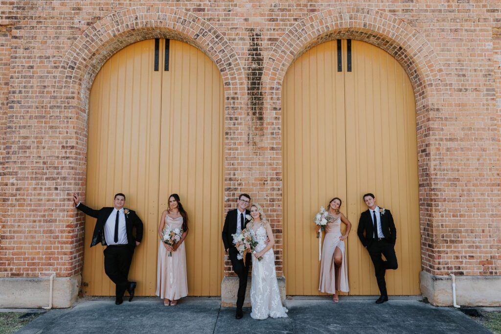 A wedding party stand in front of the large yellow barn doors of a building near Newcastle Museum. The image is fun and colourful.