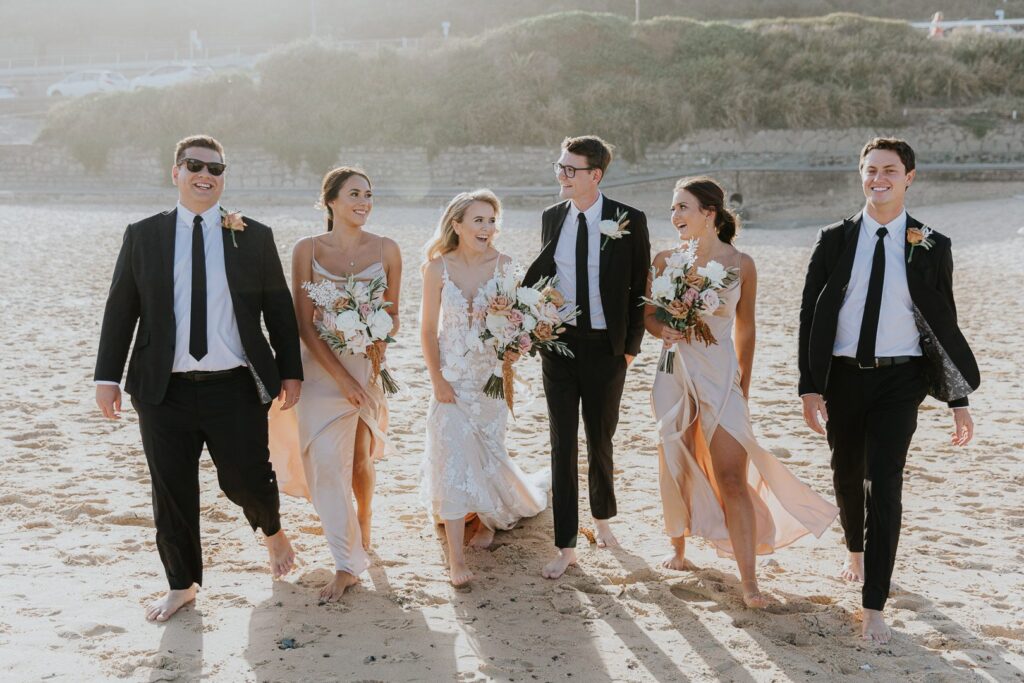 Merewether Surfhouse Wedding. A Bridal party walk down the beach laughing and having fun in the sun