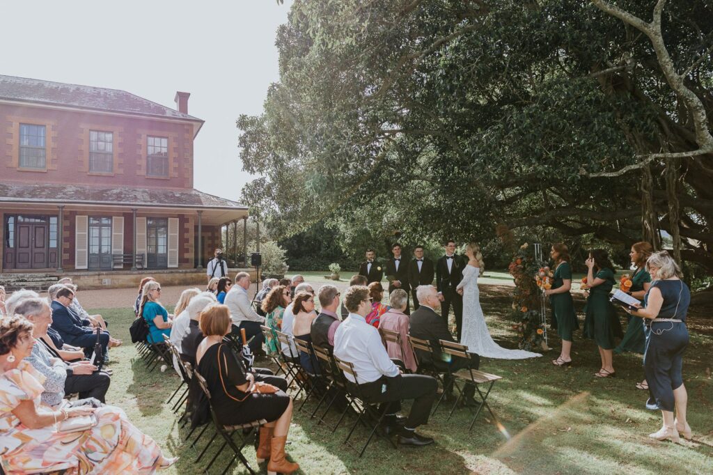 An autumn wedding at Tocal Homestead in the Hunter Valley. The couple are under a large fig tree with their guests watching on. In the background in the old Australian country manor
