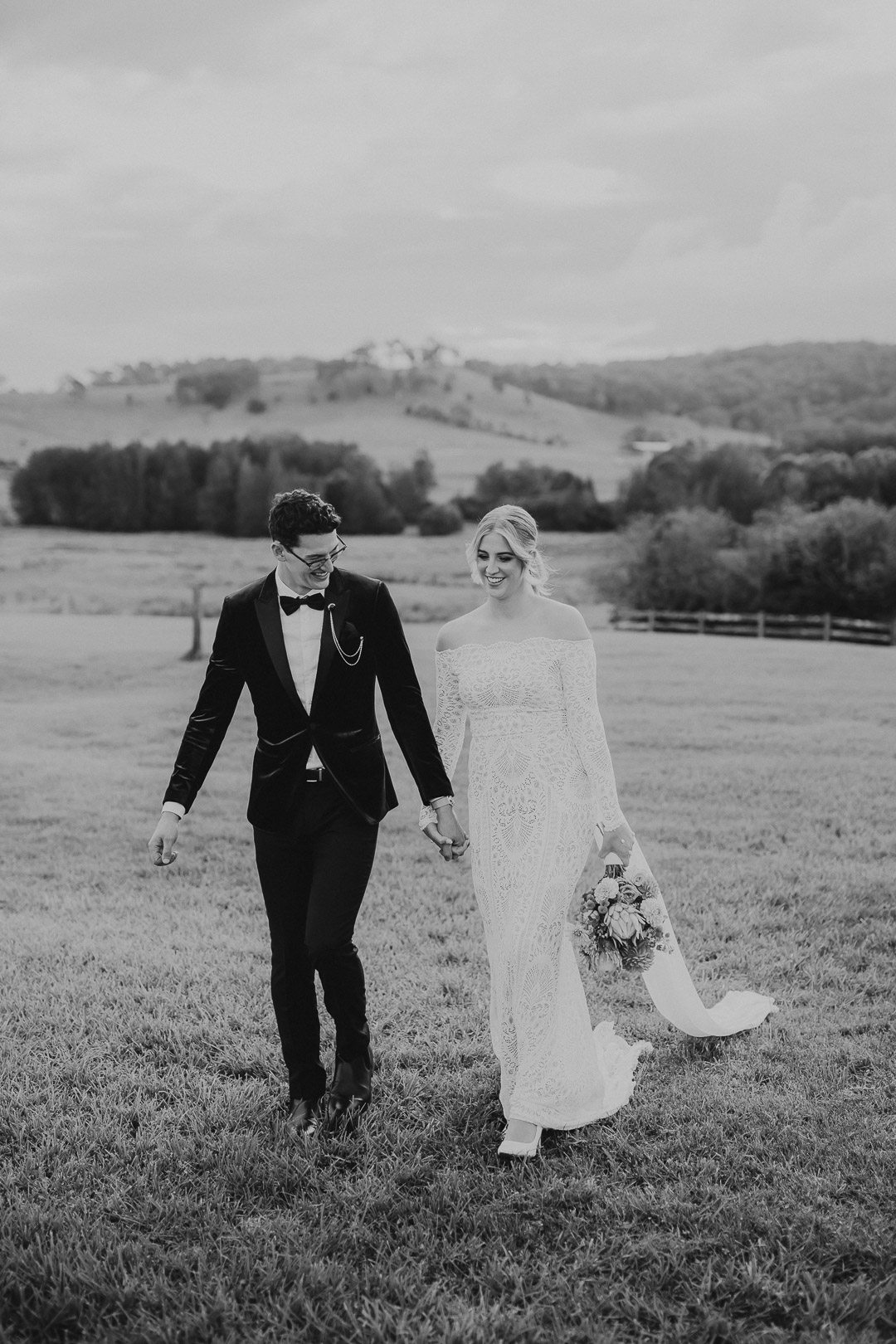 Black and white image of a bride and groom walking hand in hand through a field of grass on their wedding day. They look happy and relaxed. The image is shot at Tocal Homestead in the Hunter Valley