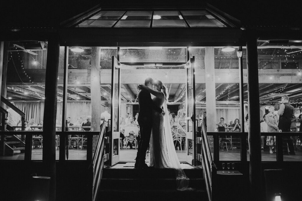 Black and white image of a wedding venue full of people, shot from outside. The venue is a wooden barn but glass fronted with windows. In the middle the bride and groom embrace each other. A very romantic and dramatic photo