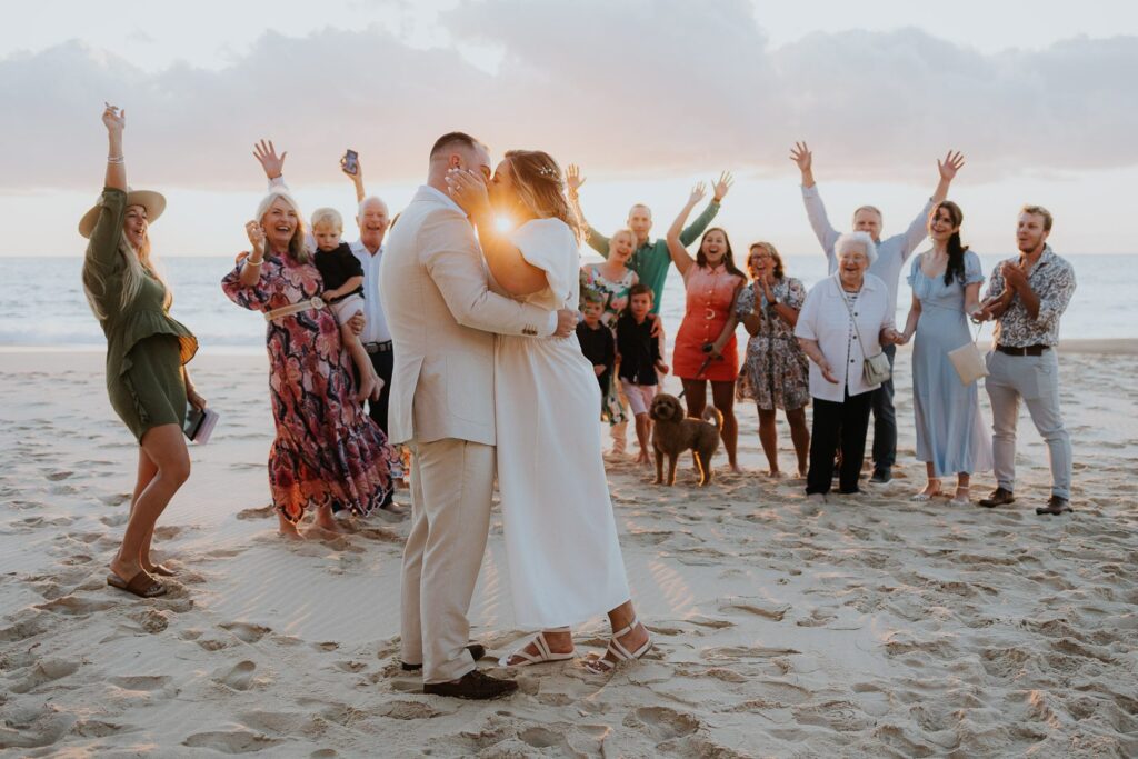 In an intimate sunrise beach Elopement in Australia, the couple kiss in the foreground, while their Celebrant and small group of family guests cheer in the background. The sun appears through the middle of their kiss. The image is fun, romantic, colourful and exciting.