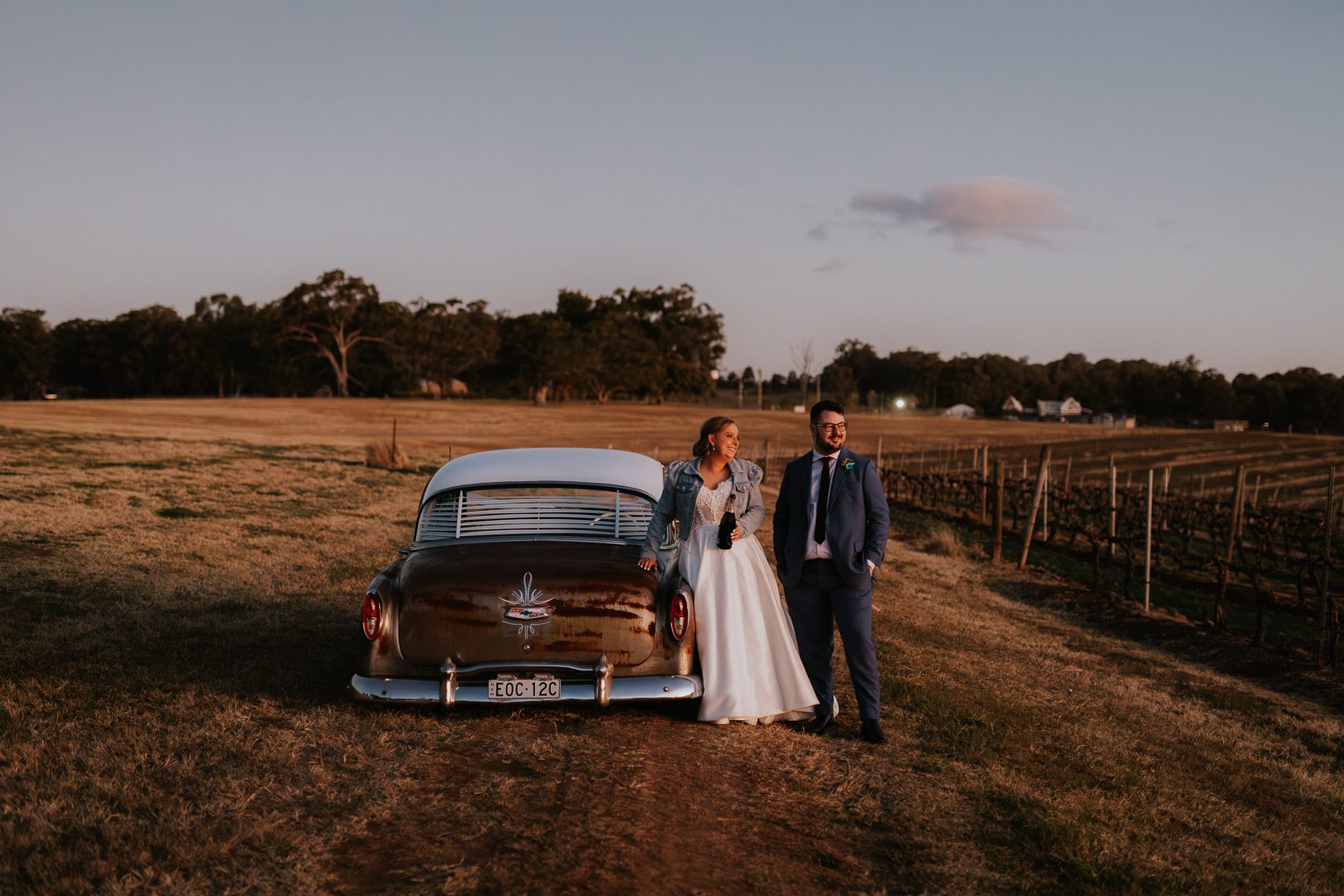 Winter Light at a wedding at Bimbadgen Palmers Lane. A couple stand beside a classic rusty american Chevrolet car. The image is warm, and they look camera right towards the setting sun as they are bathed in beautiful natural light