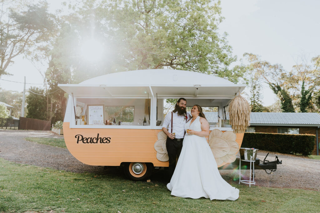 Peaches Vintage Caravan Bar at a Central Coast Wedding at The Ridge Estate, Peats Ridge, Central Coast NSW. The couple stand in front of the caravan toasting their marriage. Sunburst comes through the trees behind them.