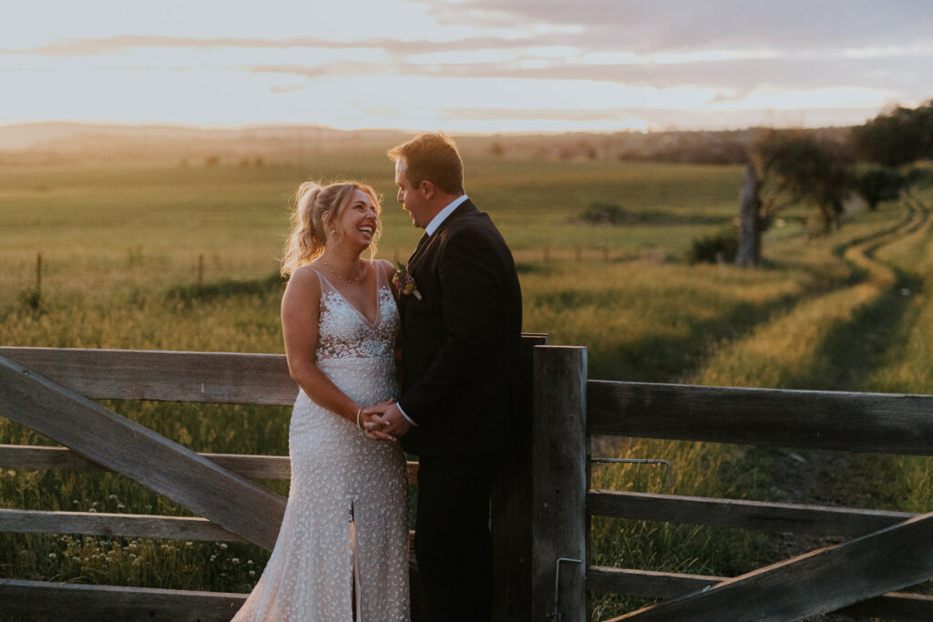 A man and woman have have just got married are holding each other and laughing, standing in front of a wooden gate with warmly lit green fields behind them. The image was shot at BoxGrove Bathurst in the Central West NSW with Underatreehouse Photography. It is a happy and relaxed wedding photo