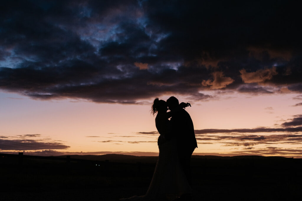 A bold and dramatic sunset wedding photo of a man and woman embracing. They are silhouetted against a yellow and purple sunset sky behind them. The image is shot from low down to fill the frame with mostly sky. The image was taken at BoxGrove Bathurst in the Central West NSW with Underatreehouse Photography.