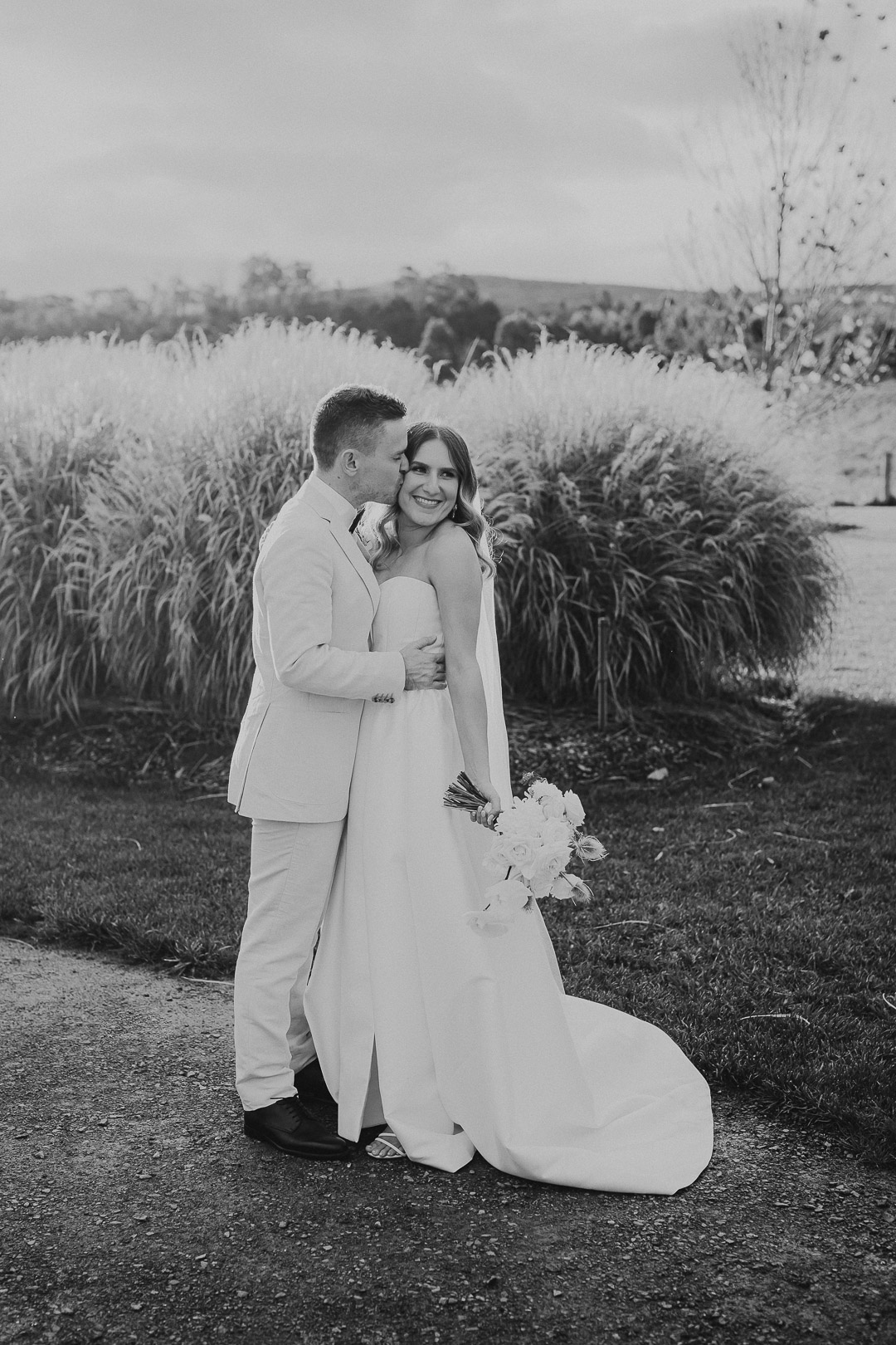 Black and white editorial style photography of a wedding at Bendooley Estate in the NSW Southern Highlands. The groom is kissing the bride on the cheek as she turns away slightly and smiles. She looks very happy. The photo is relaxed, natural and candid. Underatreehouse Photography