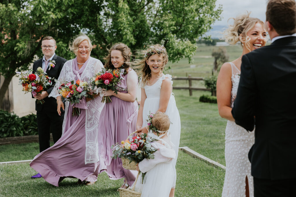 The image shows a wedding ceremony at BoxGrove Bathurst where we see the daughters and bridal party of the couple been blown away by strong cold winds. They are laughing about the fact that the wind is so strong. Its a joyous colour image.