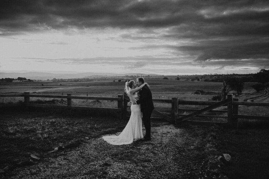 Black and white image of a wedding at Box Grove Bathurst with a dramatic sky behind the couple who are standing in front of a wooden gate kissing. The image is wide and show the rolling hills of the Central West and Bathurst behind them. Bathurst Wedding Photographer