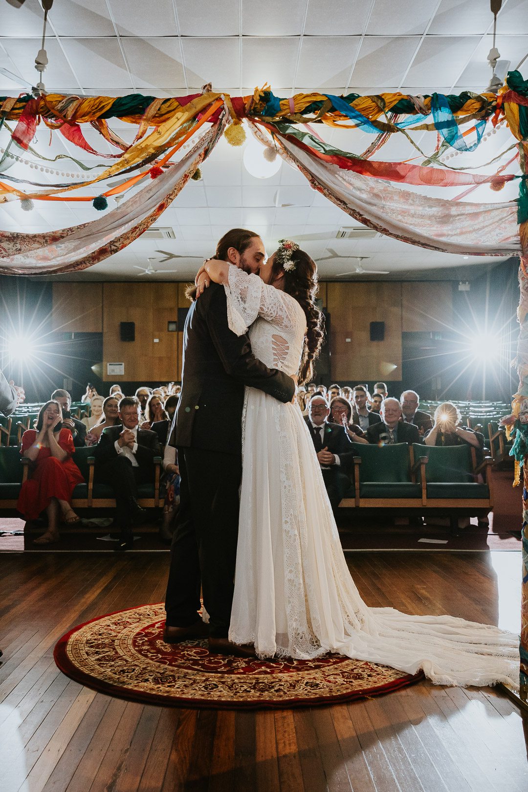 A Bride and Groom 's first kiss during their wedding ceremony. The image is shot from behind the couple, showing the guests in the background. They are framed by an arbour which is covered in colourful draped textiles. The location is a cinema at James Theatre in Dungog, Hunter Valley. Two lights at the back add pop and drama. Underatreehouse Photography