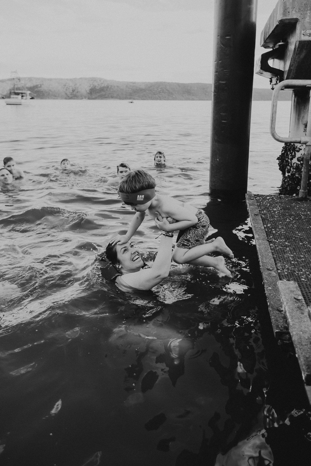 A mother is in the sea while her young son climbs off the ferry wharf onto her during their family photography session with underatreehouse photography in Patonga Beach on the NSW Central Coast