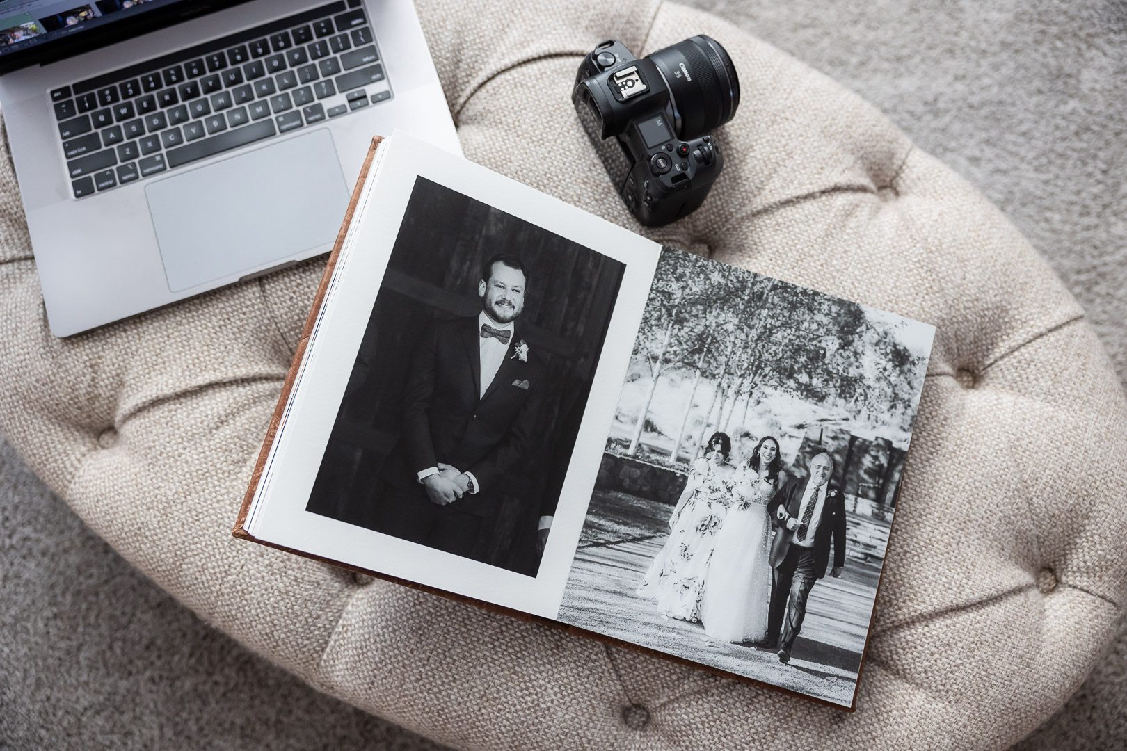 A fine art wedding album lying open on a soft furnished table with a apple laptop and canon camera lying next to it. The album shows a portrait of a groom opposite a bride approaching with her parents